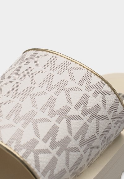 Κοντινό πλάνο λευκού λουριού σανδάλιου με επαναλαμβανόμενο μοτίβο μικρών διάτρητων γραμμάτων "MK" και χρυσή επένδυση κατά μήκος της άκρης.