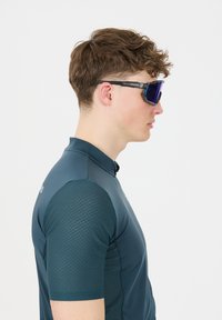 Sportsolbriller med blå spejlede linser, sorte stel og en mat finish, kombineret med en tætsiddende mørkegrønblå skjorte med teksturerede ærmer.