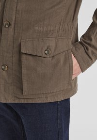 Khakifarbene Jacke mit strukturiertem Stoff, zwei vorderen Klappentaschen mit Knopfverschluss und lockerer Passform, kombiniert mit dunkelblauen Jeans.