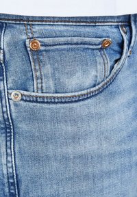 Light blue denim jeans feature a small front pocket with copper hardware and visible stitching details along the edges.