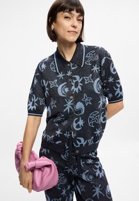 Πλεκτό μπλουζάκι με κοντά μανίκια που διαθέτει μπλε ουράνια μοτίβα σε σκούρο φόντο, συμπληρωμένο από μια ροζ τσάντα με κόμπο.