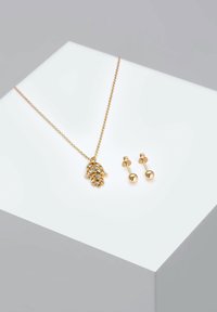Guld vedhæng halskæde med hamsa hånddesign, prydet med rhinsten; parret med gyldne kugle-stud øreringe på en hvid display.