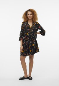 Vestido preto com estampado floral e mangas curtas em forma de sinos, apresentando um design em camadas e padrões vibrantes em amarelo, rosa e verde. Usado com sapatos planos pretos.