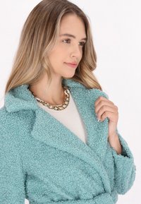 Svijetloplava, teksturirana, pahuljasta jakna s šalom, vezana u struku, i zlatni lančić preko bijele bluze. Mekan i udoban izgled.