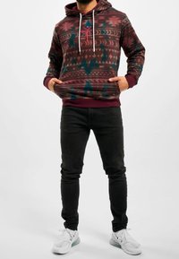 La sudadera con capucha presenta un patrón geométrico multicolor, cordones, un bolsillo frontal y puños acanalados, combinada con unos jeans negros ajustados y zapatillas blancas.