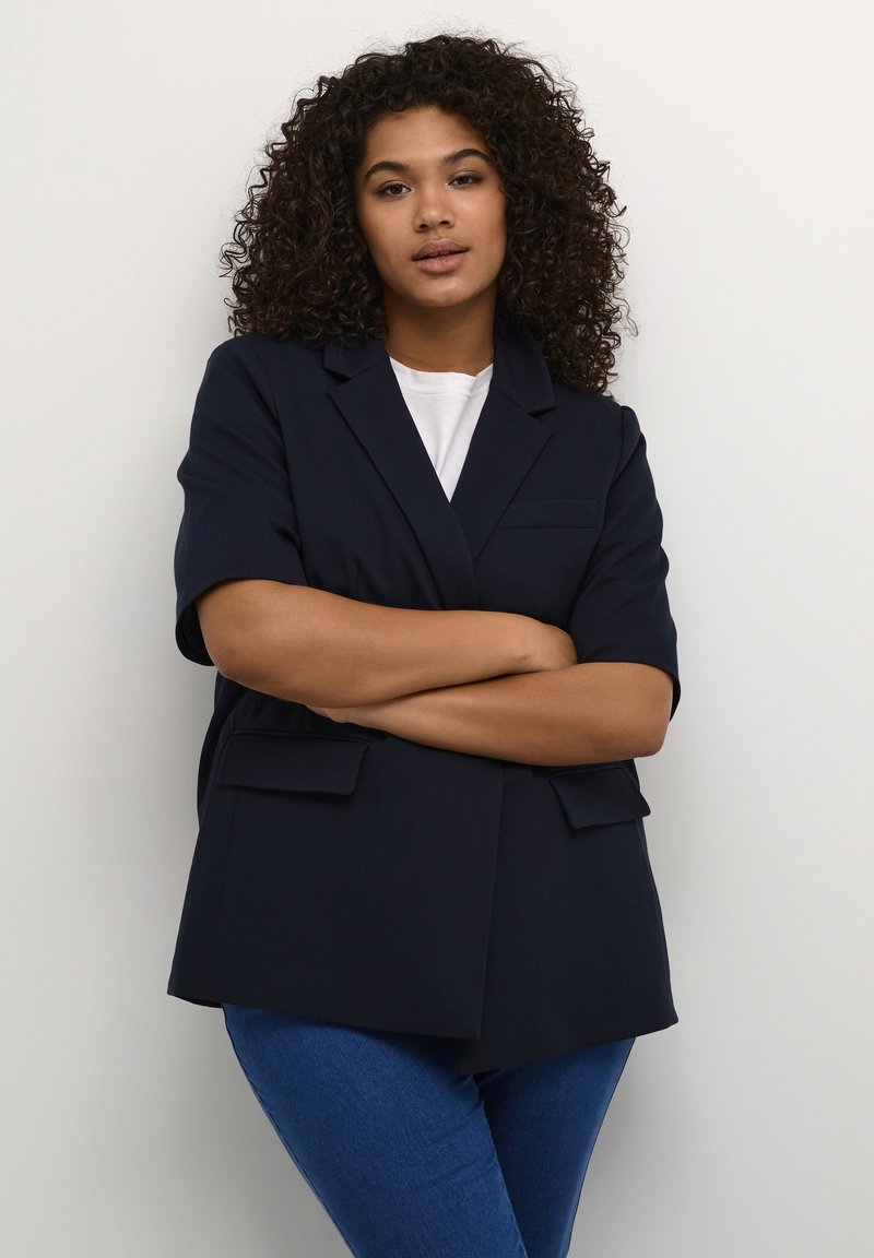 Blazer navy a maniche corte realizzato in un tessuto strutturato, con chiusura a un bottone e due tasche anteriori applicate. Indossato sopra una t-shirt bianca.