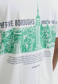 White cotton t-shirt featuring green skyline graphic of New York City with text "FIVE BOROUGHS HIDDEN IN THE HEIGHTS" and brand name below.