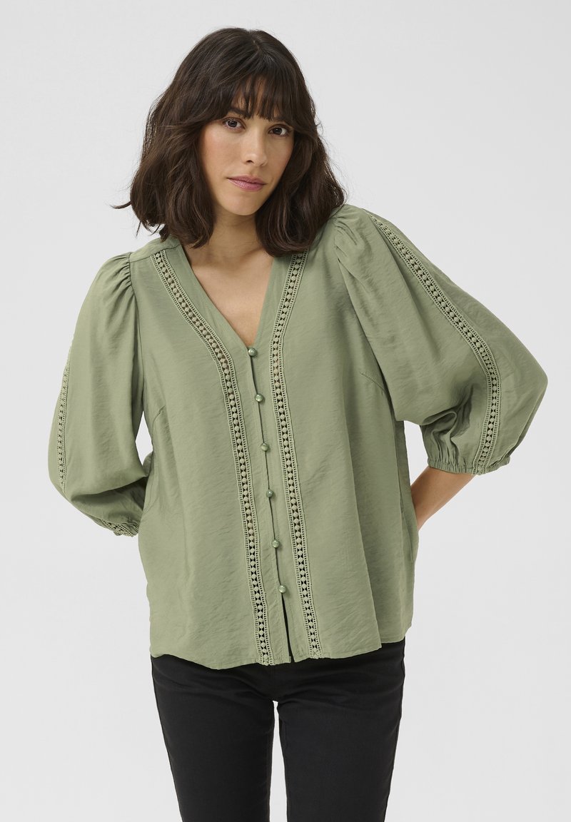 Blusa verde, leggera, con scollatura a V, maniche a palloncino e rifinitura in pizzo decorativo sul davanti. Presenta chiusure con bottoni e una vestibilità rilassata.