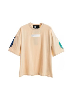 Joven de pie, lleva una camiseta beige holgada y pantalones cortos con detalle de cadena, calcetines blancos con texto y zapatos beige texturizados sin cordones.