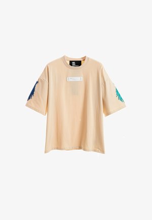 Joven de pie, lleva una camiseta beige holgada y pantalones cortos con detalle de cadena, calcetines blancos con texto y zapatos beige texturizados sin cordones.