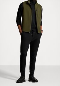 Olivgrön zip-up väst över en svart långärmad tröja, kombinerat med svarta sweatpants och svarta träningsskor. Har en ficka med dragkedja på västen.