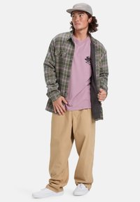 Grünes und violettes kariertes Flanellhemd über einem hellrosa T-Shirt; beige locker sitzende Hose; graue Kappe; weiße Turnschuhe.