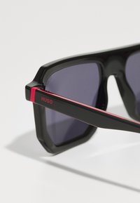 Ochelari de soare negri cu ramă pătrată, având lentile întunecate. Accente roșii pe montant cu logo-ul "HUGO". Finisaj lucios și design îndrăzneț.