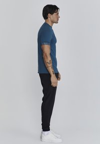 Camiseta de algodón azul con mangas cortas, que presenta un logo en el brazo, combinada con pantalones jogger negros y zapatillas blancas.