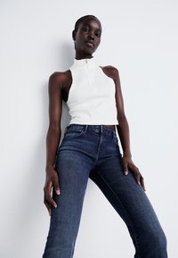 Haut sans manches blanc à col montant et fermeture éclair, associé à un jean fitted bleu foncé. Le modèle se tient avec les mains sur les hanches devant un fond clair.
