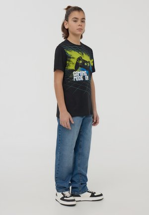 T-shirt nera a maniche corte con grafica di un controller di gioco e testo "Modalità Gioco Attivata", abbinata a jeans azzurri chiari e sneakers bianco e nero.