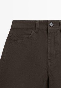 Donkerbruine broek met hoge taille, met knoopsluiting aan de voorkant, broekriemlussen en een voorzak, weergegeven van taille tot bovenbeen.