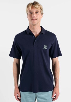 Homme blond portant un polo à manches courtes bleu marine avec un petit logo coloré sur le côté gauche de la poitrine et un pantalon bleu clair.