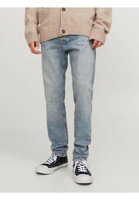 Persona che indossa jeans azzurri chiari, cardigan beige e sneaker bianco e nero, in piedi contro uno sfondo chiaro e semplice.