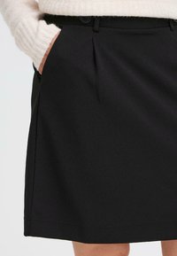 Zwarte plissé rok met een gladde textuur, een tailleband met twee knopen en schuine zijzakken. Zichtbare stiksels langs de zoom.