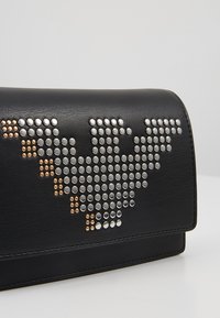 Černá kožená kabelka s kovovými hroty ve stříbrné a bronzové barvě uspořádanými do geometrického vzoru na předním klopě.