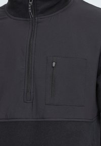 Schwarzer Pullover mit hohem Kragen, halbem Reißverschluss vorne und einer Reißverschlusstasche auf der Brust. Verfügt über einen glatten Stoff und einen strukturierten Fleece-Unterbereich.