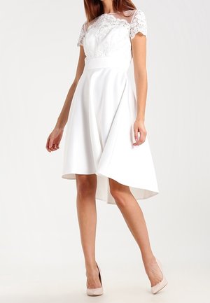 Vrouw die een witte jurk tot op de knie draagt met korte kanten mouwen en een asymmetrische zoom, gecombineerd met beige hoge hakken.