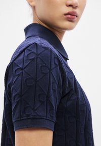 Tumši zils polo krekls ar teksturētu rakstu, kurā redzamas savstarpēji savienotas ģeometriskas formas. Īsas piedurknes un klasisks apkaklītis ar cieši austu apdari.