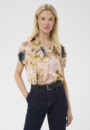 Vrouw die een bloes met korte mouwen en bloemmotief draagt, ingestopt in een donkerblauwe spijkerbroek met een zwarte riem, staand met één hand in haar zak.