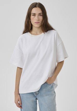 Fiatal nő hosszú barna hajjal, túlságosan nagy fehér pólót és világoskék farmert visel, egyik kezét a zsebébe dugva áll, egyszerű háttér előtt.