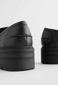 Black platform shoes with a textured leather upper, rounded toe, and contrasting sole grooves. Features a low-cut design.
