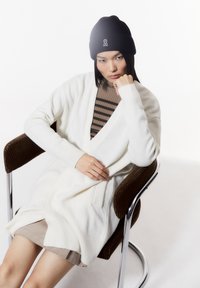 Weißer, offener Cardigan aus weichem Material mit langen Ärmeln, getragen über einem braun gestreiften Oberteil, sitzend auf einem modernen Stuhl mit Chrombasis.