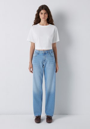 Young woman with long curly hair wearing a plain white short-sleeve crop top, light blue high-waisted jeans, and brown shoes standing.