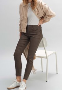 Brun slim-fit housut, joissa on vetoketjullinen tasku; yhdistetty beigeen corduroy takkiin ja valkoisiin tennareihin. Yksinkertainen, räätälöity muotoilu.