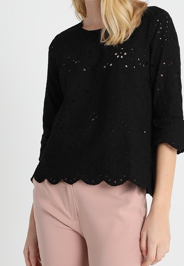 Femme portant un chemisier en dentelle noire ajourée avec ourlet festonné et manches trois-quarts, associé à un pantalon rose clair.