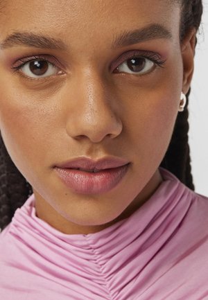 Acercamiento de un rostro con piel de tono marrón medio, ojos marrones, sombra de ojos sutil y labios rosa suaves; vistiendo una blusa de color rosa claro, texturizada y con pliegues.