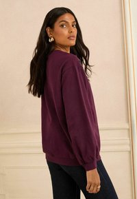 Violet sweatshirt su didelėmis rankovėmis ir briaunotais rankogaliais, derintas su tamsiomis džinsinėmis kelnėmis, prieš neutralų foną. Matomas subtilus prekės ženklo detalė.