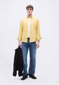 Νεαρός άνδρας όρθιος, φοράει κίτρινο πουκάμισο, λευκό T-shirt, μπλε τζιν, μαύρα παπούτσια, κρατώντας ένα μαύρο σακάκι στο αριστερό του χέρι, απλό λευκό φόντο.