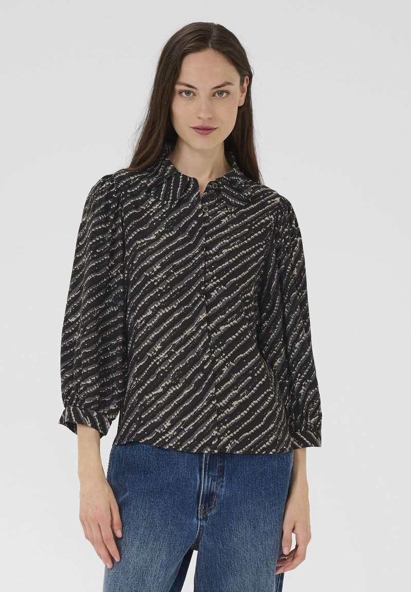 Egy nő fekete és bézs átlós csíkos blúzt visel puffos ujjal és kék farmernadrággal, egy sima fehér háttér előtt áll.