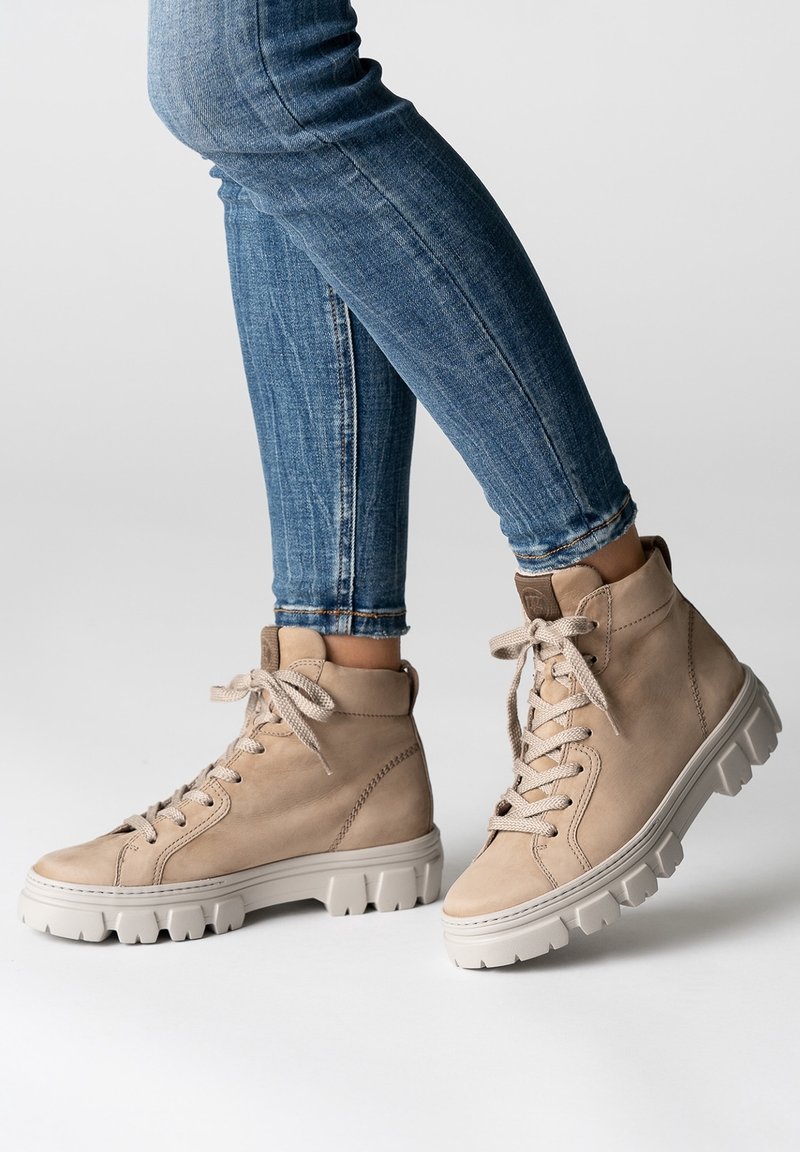 Stivali alti in pelle beige con suole bianche spesse indossati con jeans skinny blu, mostrati su uno sfondo bianco.