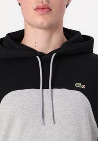 Juoda ir pilka kapučino tipo megztinis, turintis kengūros kišenę, pilkas virvutes ir mažą žalią Lacoste krokodilo logotipą ant krūtinės.
