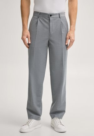 Uomo che indossa pantaloni eleganti plissettati grigio chiaro, camicia bianca infilata e sneakers bianche, in piedi rivolto in avanti contro uno sfondo neutro.