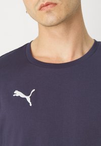 Mørkeblå t-skjorte laget av glatt stoff, med en hvit brodert Puma-logo på venstre bryst, og med en klassisk rund halsdesign.