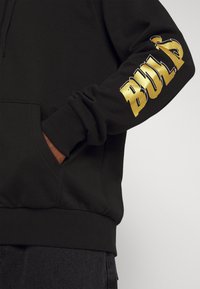 Sudadera negra con puños y dobladillo de canalé, que presenta un gran gráfico dorado "BULD" en la manga izquierda. Bolsillo tipo canguro en la parte frontal.