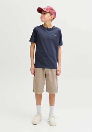 Dreng iført lyserød kasket, marineblå t-shirt, beige shorts, hvide sokker og sneakers, står og kigger til siden mod en ensfarvet baggrund.