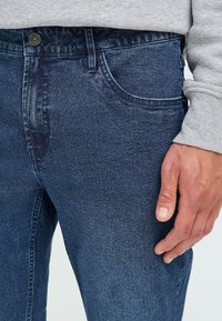 Blugi din denim de culoare albastru închis, cu o croială clasică. Au cinci buzunare, închidere cu buton și detalii discrete de cusătură de-a lungul cusăturilor.