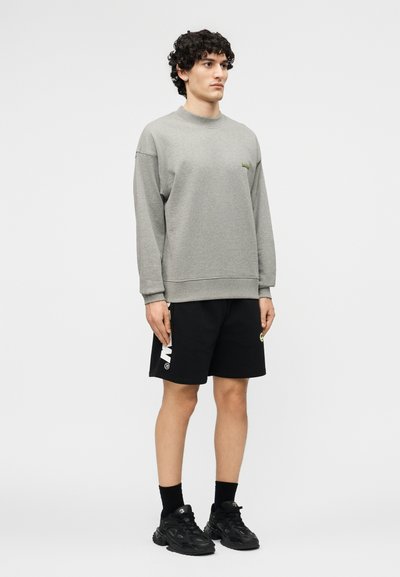 Νεαρός άνδρας που στέκεται, φορά γκρι φούτερ με στρογγυλό λαιμό, μαύρο σορτς με λευκό λογότυπο, μαύρες κάλτσες και μαύρα αθλητικά παπούτσια, μπροστά σε απλό φωτεινό φόντο.