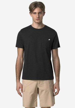 Giovane uomo che indossa una semplice t-shirt nera a maniche corte con una piccola tasca sul petto e pantaloncini cargo beige su uno sfondo neutro.