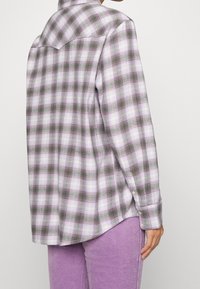 Chemise en flanelle à carreaux violets et verts avec un col classique, des détails de boutons et une coupe décontractée, portée avec un pantalon en velours côtelé violet.
