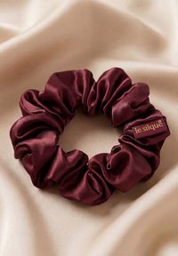 Donkerrode satijnen scrunchie met een klein "le sique" label rustend op zachte beige stof.